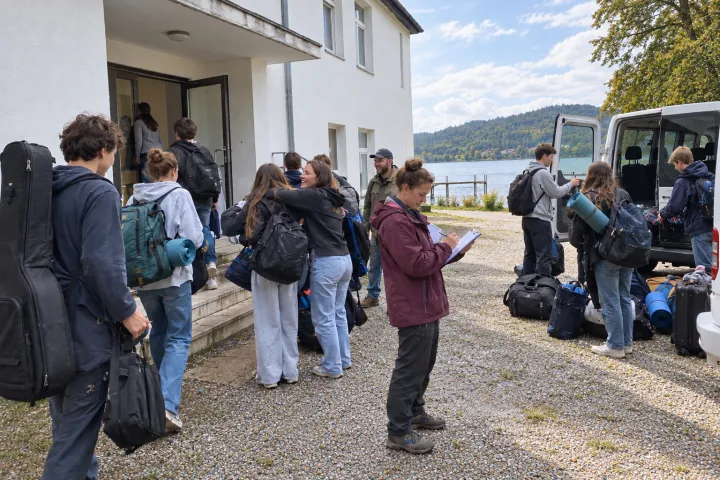 Jugendliche mit Rucksäcken, Isomatten und einem Gitarrenkoffer kommen an einem weissen Freizeithaus am See an; eine Betreuerin hakt auf einem Klemmbrett ab, hinten wird ein Kleinbus ausgeladen.